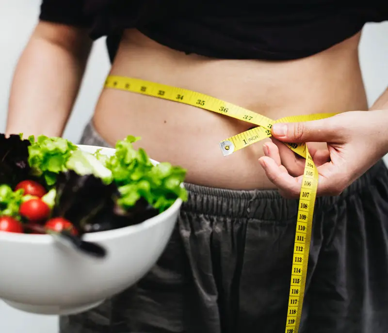 A woman is measuring her waist with a bowl of salad.