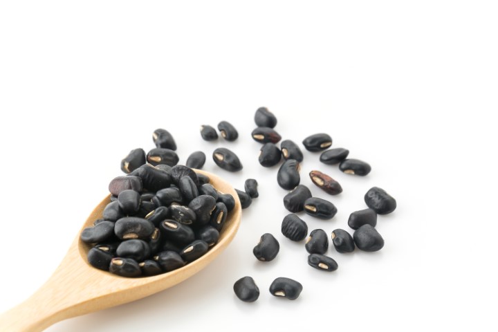 A wooden spoon filled with black beans on top of white surface.